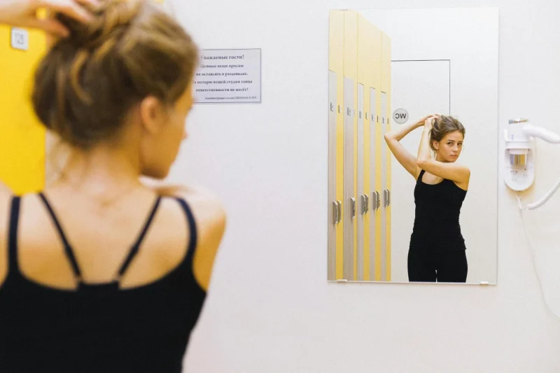 Žena u sportskoj majici vezuje kosu ispred ogledala u svlačionici.